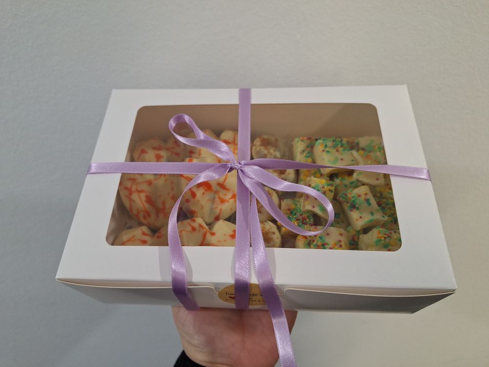 Eine Box mit lila Schleife. Zu sehen sind weiße Herzen mit orangenen Sprenklern, Cakepops mit Biscoff Krümel und helles Fudge mit bunten Streuseln. 
Gehalten wird die Box von einer Hand. 