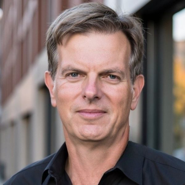 Headshot of a middle-aged white man with short, light brown hair and blue eyes. He wears a black collared shirt. The background is blurry, appearing to be an outdoor urban setting with brick and glass.