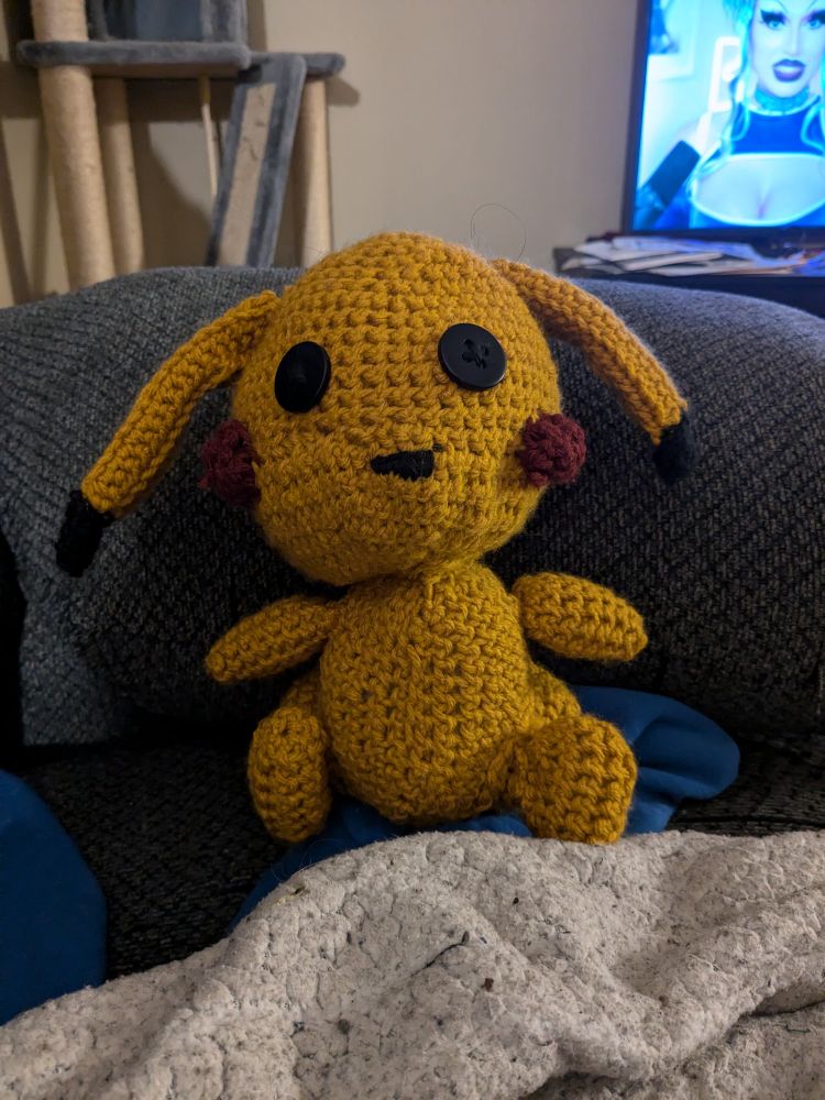 A crochet Pikachu plushie that is sitting down, with black button eyes. He's a little wonky and lopsided, but has that vintage Winnie the Pooh vibe 