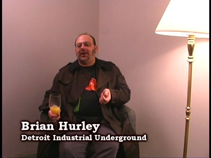A man wearing a trench coat over a t-shirt sits in a low chair in a bare, beige corner lit by a floor lamp. He is holding an orange drink in a wine glass in one hand and gesticulating with his other hand. Text in the lower third reads: Brian Hurley, Detroit Industial Underground.