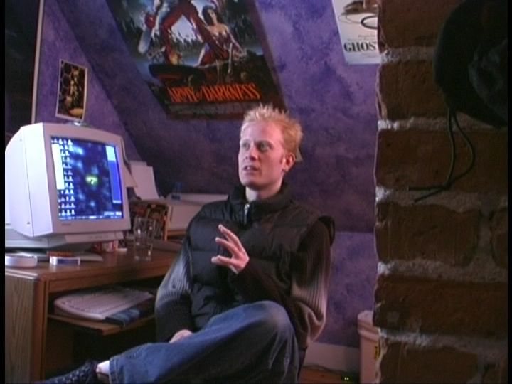 A man sits at a PC setup tucked away in the corner of a room with a low, sloped ceiling. A CRT monitor sits atop a desk with a pull-out keyboard tray. Posters for Ghost in the Shell and Army of Darkness are pinned to the ceiling.