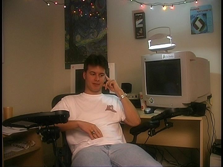 A man sits in an office chair in front of a computer desk. The armrests of the chair have extensions to hold keyboards and space to use a mouse. The desk in the background has two CRT monitors. Christmas lights are strung about the ceiling.