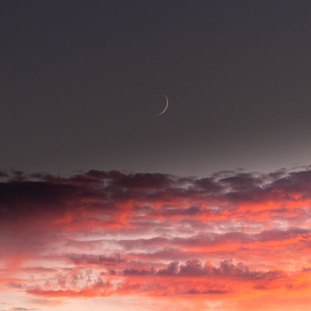 A thin crescent moon in the sky over some cloud catching the sunset rays.