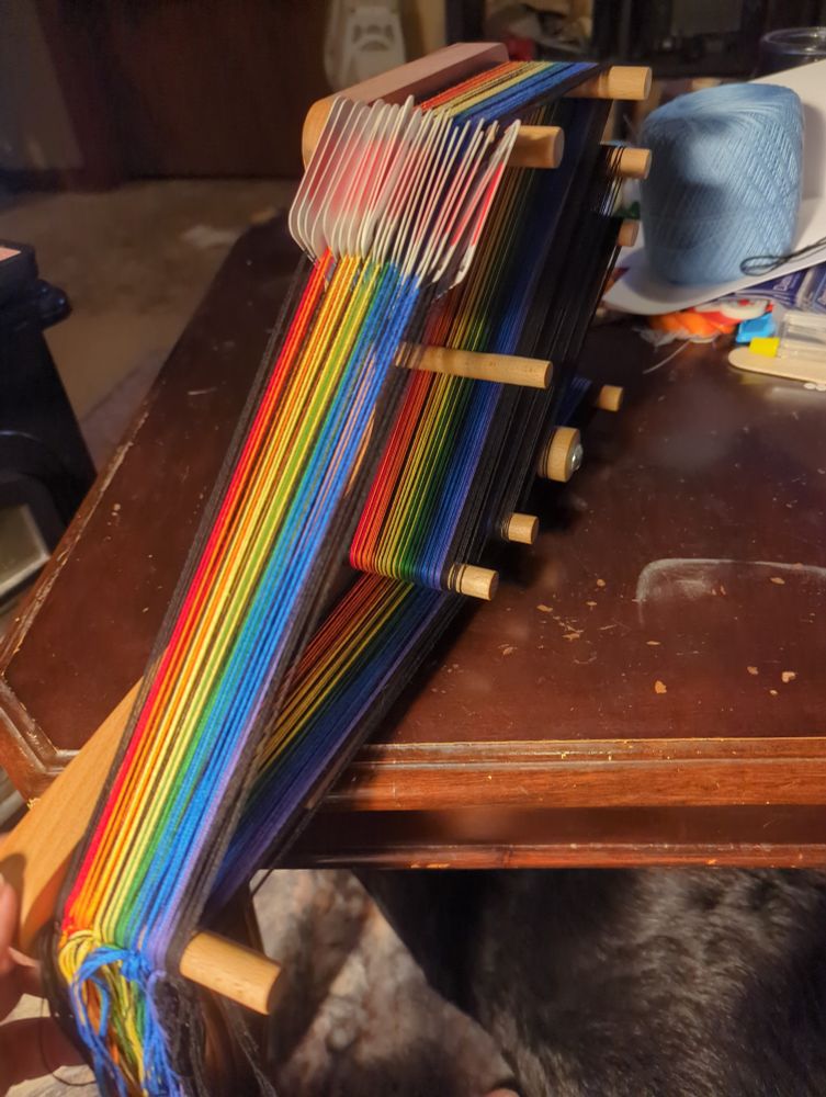 An inklette loom warped with a rainbow of threads sitting on a coffee table.  The warp is run through cards for tablet weaving. 