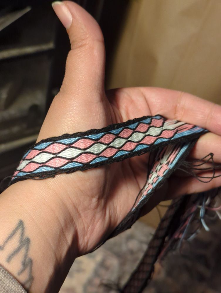 Imriel's hand is visible with a tablet woven trans flag colored band around their wrist. They are pinching the ends together with their fingers to show the design.