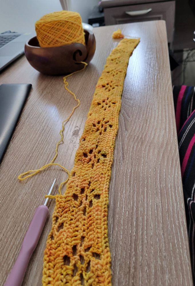 A long, thin line of crochet stitches displaying a floral lace pattern. The yarn is a warm golden yellow with subtle red and green tones blended in. A lavender crochet hook is attached, and the yarn cake is in a wooden bowl 