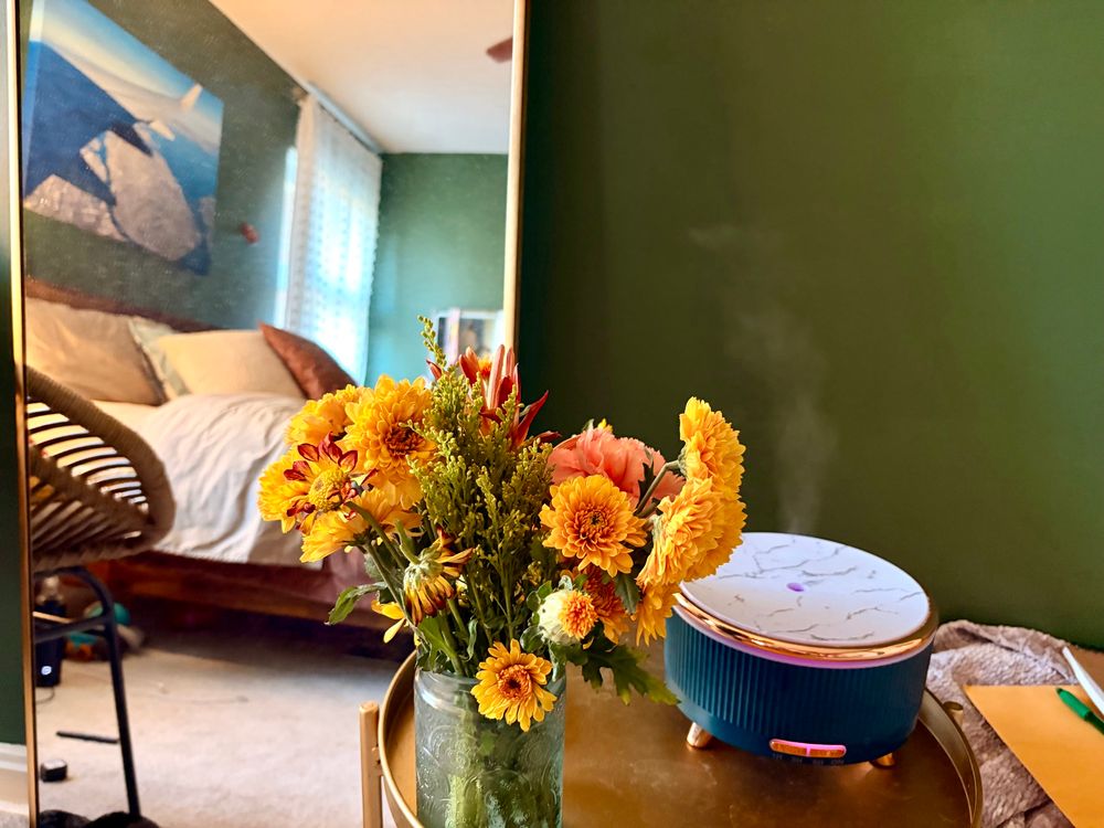 A bouquet on a gold circular tray with a green modern oil diffuser behind it on the table. There’s a rectangle gold rimmed mirror leaning against the wall. In the mirror, you can see a dark green wall and a bed that is made. There is a photo taken from an airplane of the San Francisco Bay. 