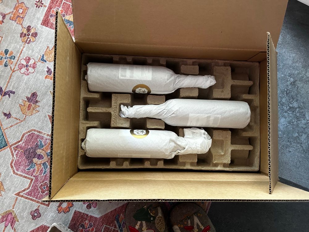 A photo of an open cardboard box. It’s three bottles wrapped with a gold logo showing. The wine is As Ever