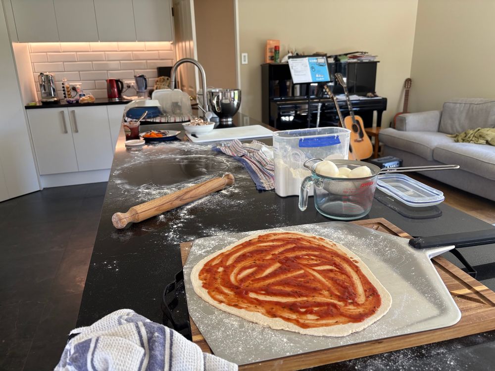 A pizza base on a peel with no toppings. Rolling pin, flour, and mozzarella on the kitchen side showing pizza making in progress.