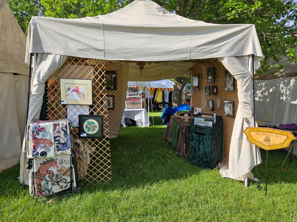 Ezyup style tent covered in raw canvas. Open in front and back. On the left front is a lattice holding two pieces of art - one showing a red dragon coming out of the page, the lower a round Celtic style piece of three horses in red, blue, and green. Also on the left is a rack with several folded blankets.