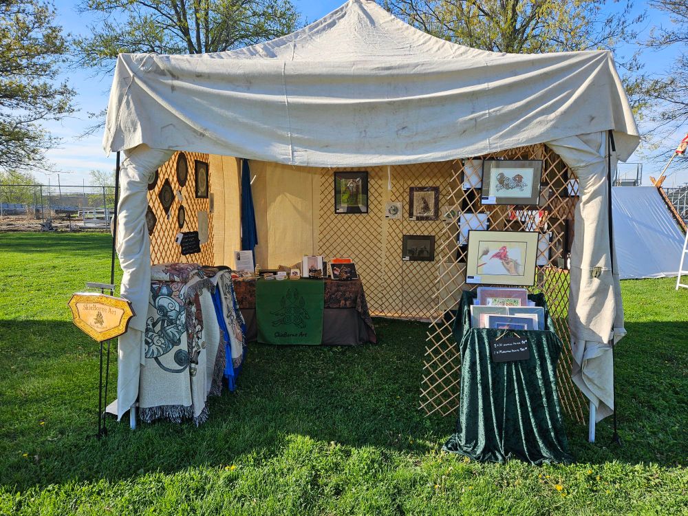 Pop up tent covered in cream canvas with artwork on hanging trellis inside. Prints in a bin on the right and blankets to the left.