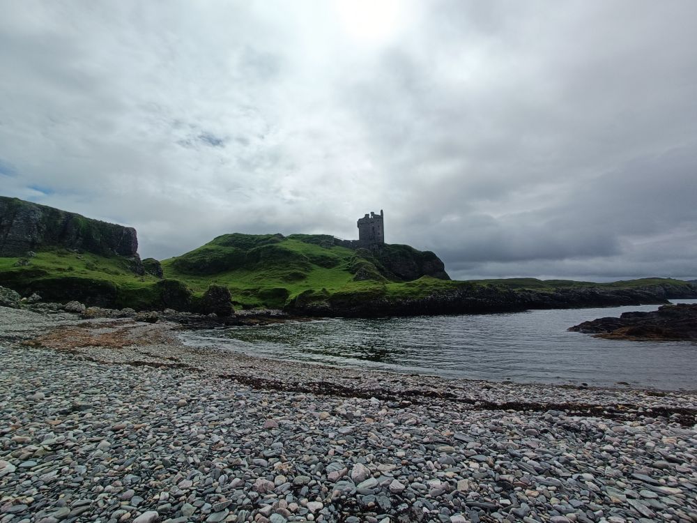 Gylen Castle on Kerrera, standing on a hill with a pebbly beach and calm sea in front, light clouds above.