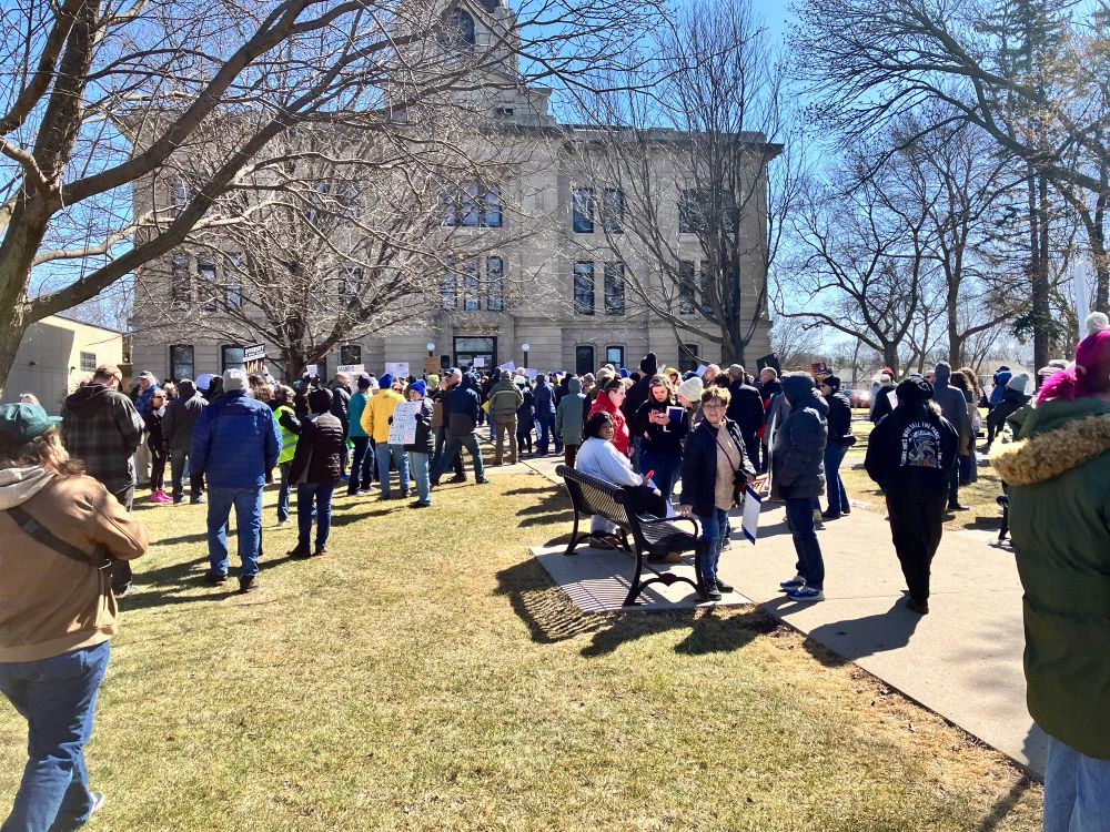 A crowd of people at a protest in front of a courthouse. The grass is showing early signs of spring green, but the trees are still bare.