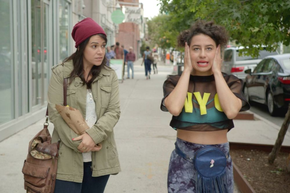Abby and Ilana from Broad City walking down a NYC sidewalk. Ilana makes a goofy expression while holding her head in her hands, wearing a mesh NYC crop top, while Abby looks on.