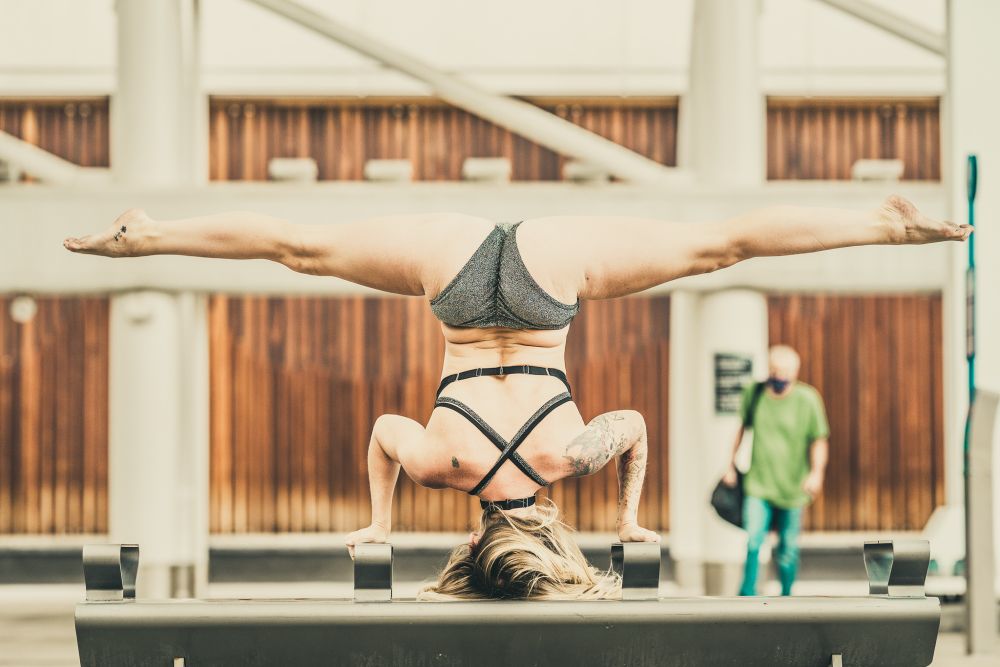 A blonde yoga girl in her black underwear does a headstand and splits on a bench at a train station.