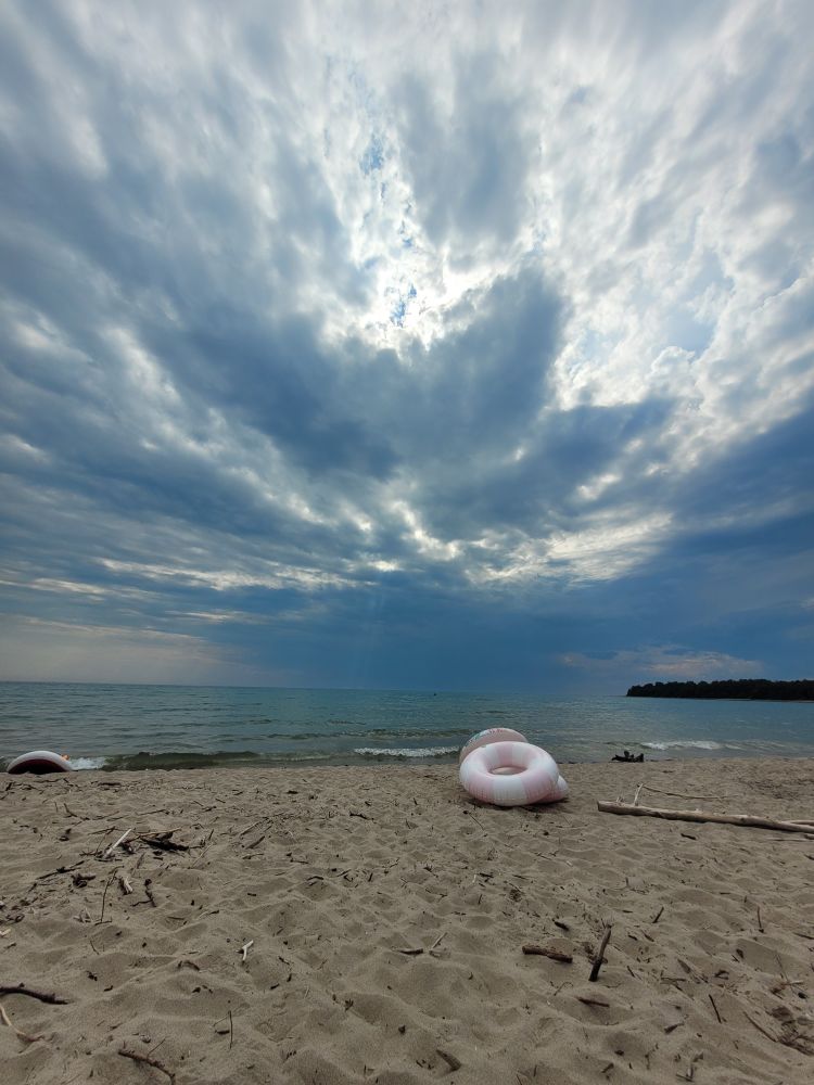 A photo of the shoreline, beach sand in the foreground, with a stack of inflatables. The sky is clouded but sun shines through the cracks. At the horizon, there is a smudge of rain.