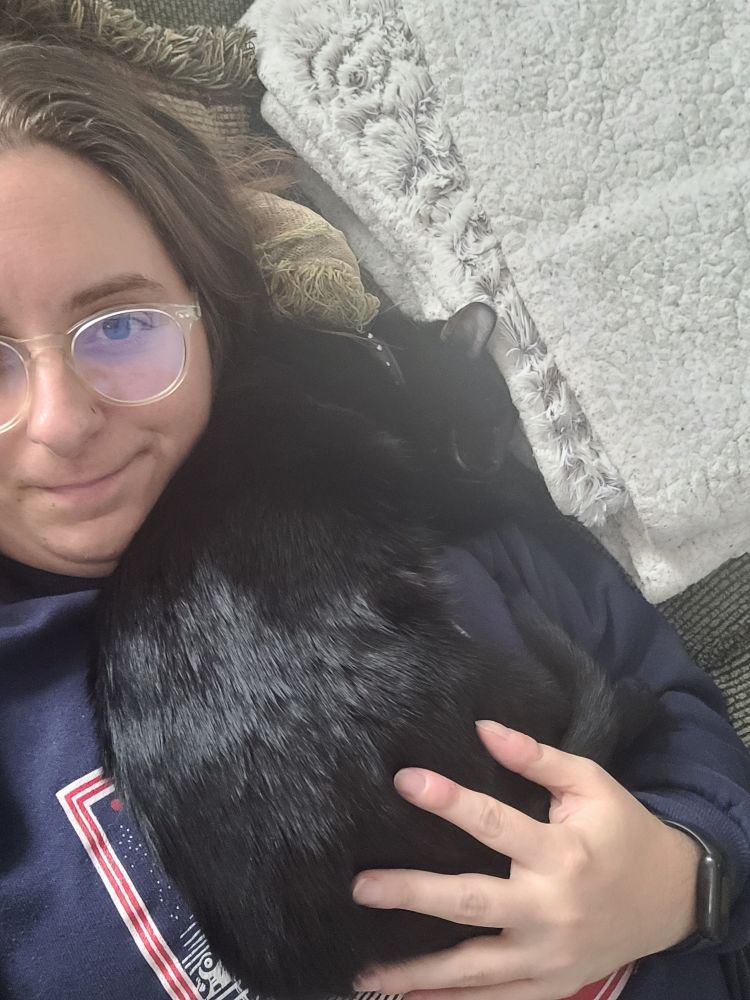 A selfie of me holding the black cat. He is curled up on my chest. I am wearing glasses and half-smiling.