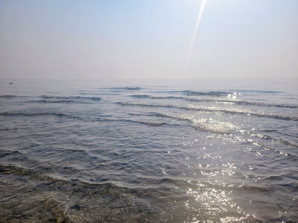 A photo of the view looking out over the water. The surface is wavy and dappled with sun. The horizon is undefined as the sky has a haze of wildfire smoke.