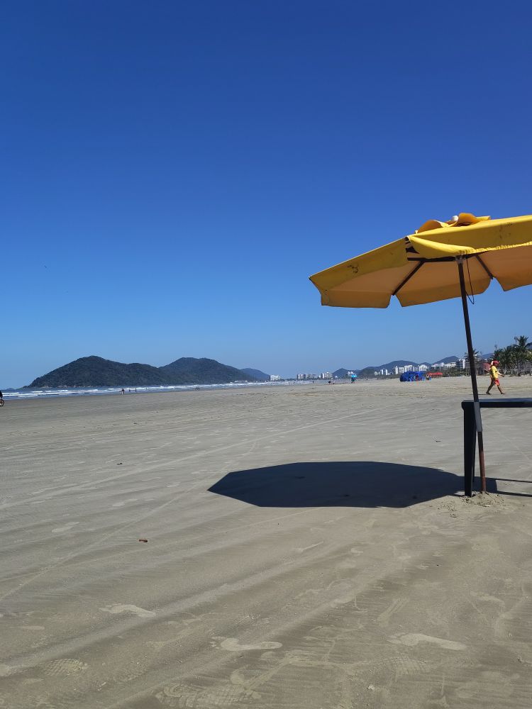 Praia, céu e sol de Bertioga com ombrelone amarelo e mesa de plástico. 