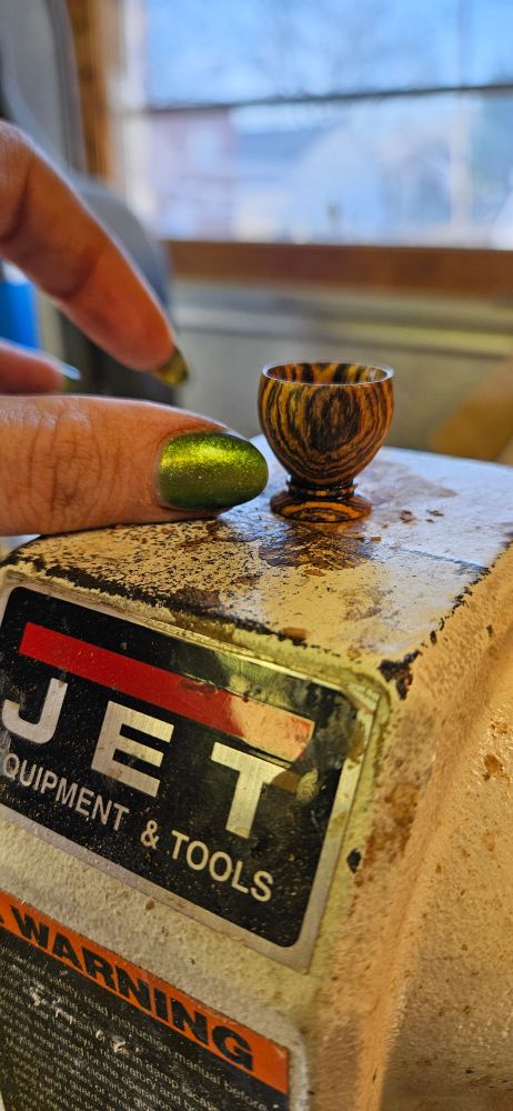 Side profile of a miniature woodturning that bri made from Bocote wood which is yellow with black/brown grain lines. Her thumb is next to the form for scale. The finished object, which could be called a bowl with a decorative base? is approximately 1.5 thumbs tall and approximately the same diameter. 