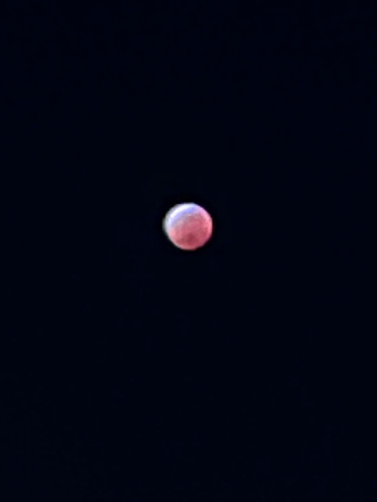 An amateur photo of the lunar eclipse. The moon is a little blurry but you can just make out darker patches. The moon is mostly glowing red with a soft white crescent on the top 