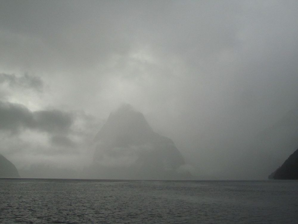 A shadow of an island appears indistinct through a veil of heavy clouds and smog, rising from an expanse of open, grey water.