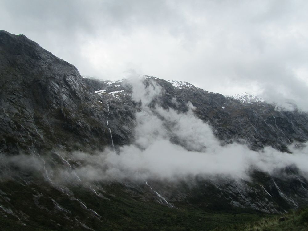 Dark mountains dominate the image, its snowcapped peaks touching the overcast sky. Thin waterfalls trace down from peaks to the valley.