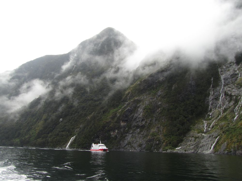 A white ship with a red hull is dwarfed by majestic mountains enriched in green vegetation, little ribbons of waterfalls streaming from peak to the open water reflecting the mountains.