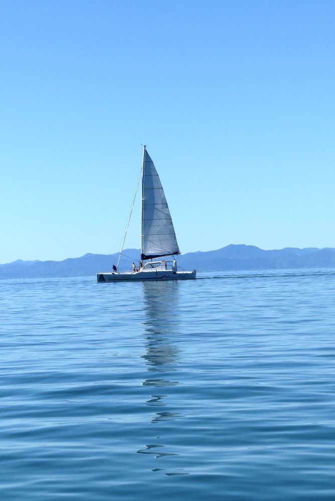 A boat with a tall white sail and body sails on calm ocean water, its reflection clear in the ripples. Mountains, appearing blue, rise a little into the cloudless blue sky.