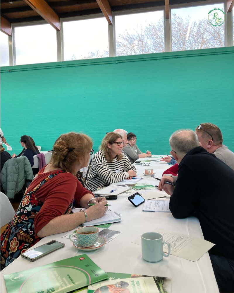 People seated around a table in discussion, taking notes and looking at printed documents and booklets. The room has tall windows and bright turquoise walls.
