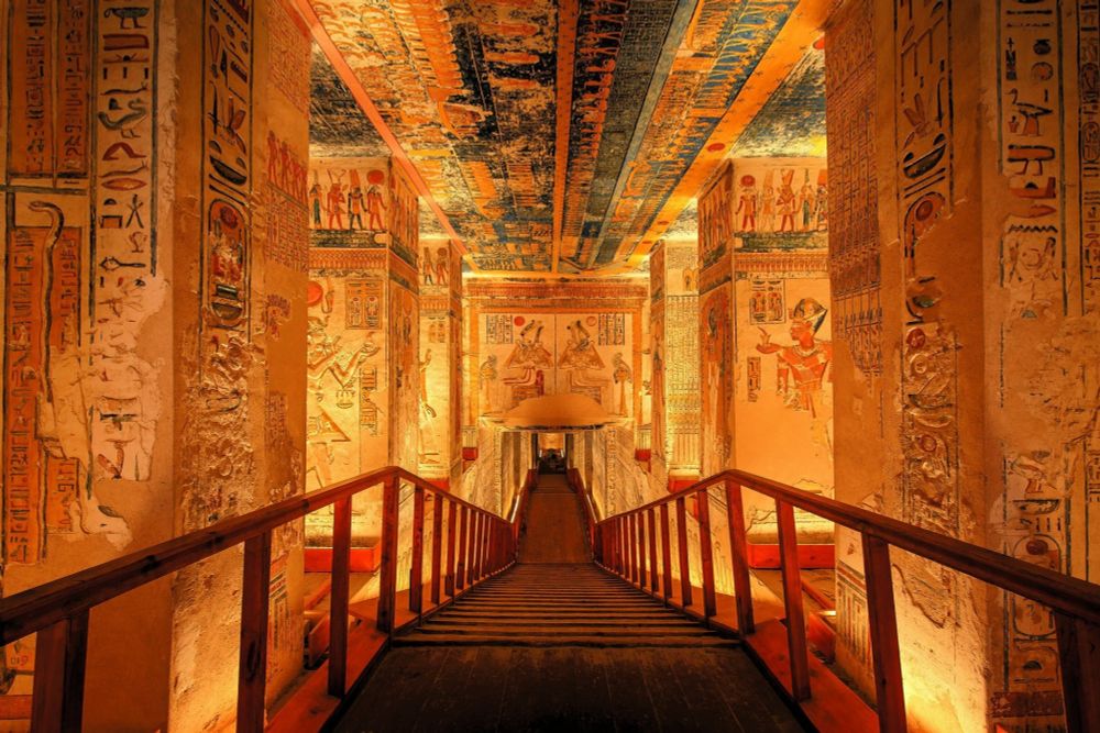 Photo of the inside of an Ancient Egyptian tomb where numerous wide stone pillars are covered with colourful hieroglyphics and writings. It's a vision of warm colors of amber, red, gold, brown, and blue.