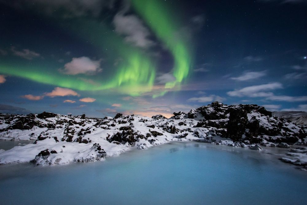 Cool green shimmers of the Northern Lights or Aurora Borealis streak across the sky above a lagoon of turquoise blue water surrounded by snow-covered black rocks. The lagoon is the famous Blue Lagoon in Iceland. 