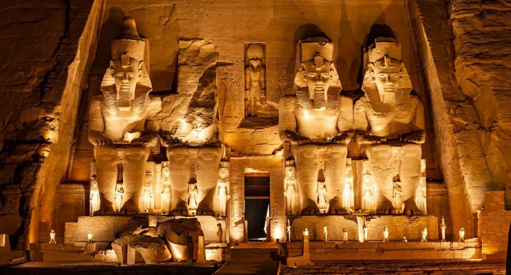 The four colossal statues of Ramses II at Abu Simbel gleam in the side of rock from which they are carved. Three of the figures are still fully intact. All four are seated figures that are lit up by discreet recessed lightinh so that the towering ancient Egyptian figures gleam like gold against the night sky. Humans are tiny figures against the statues' colossal imposing height. Two statues are on each side of a small door in the center that leads inside the temple complex. 