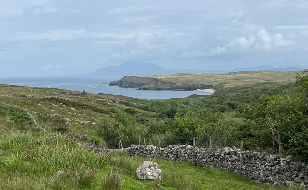 Green fields with a stone wall and some stone ruins. Beyond them are some cliffs overlooking a bay with a mountain off in the distance beyond in the mist. 