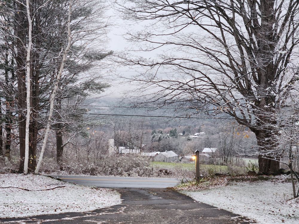 Wintry New England scene—driveway flanked with snowy yard, lamppost at the bottom of the driveway, mid-ground trees framing a view of a farm across a snow-frosted green field, mountains in the distance.