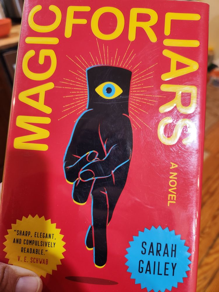 Cover art for Magic For Liars, by Sarah Gailey: bright red background, upside down, shadowy human hand with fingers crossed and an eye graphic—yellow with light blue iris—on the wrist.
