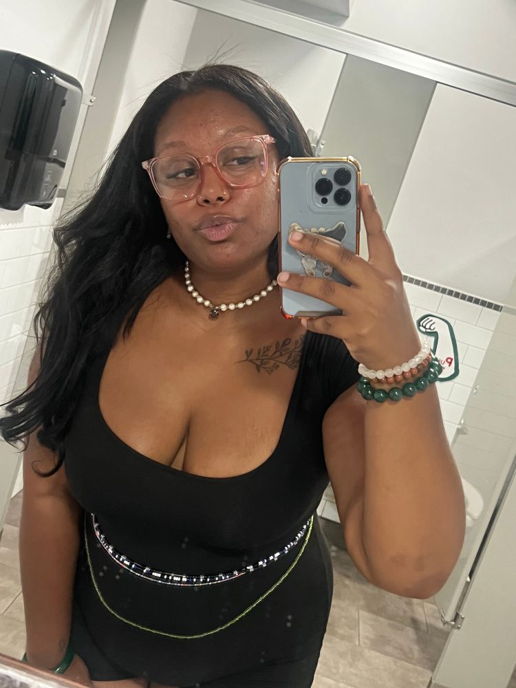 Princess in an all black shorts romper leaning forward into the mirror, making a slight kissy face and holing there phone up to take a picture 