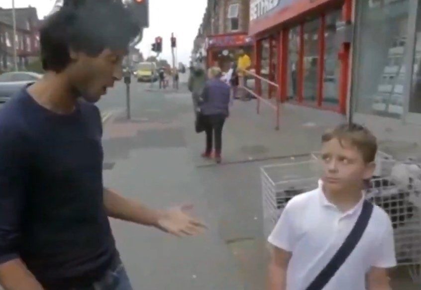 Cena em vídeo de rua, filmada em uma calçada urbana. À esquerda, o ator Assad Zaman (um homem jovem, de cabelo escuro e camiseta escura) gesticula com uma das mãos enquanto fala de forma exaltada. À direita, um menino de camiseta branca e bolsa transversal olha para ele com expressão desconfiada e confusa. Ao fundo, há lojas com fachadas vermelhas, pedestres caminhando e um cruzamento com semáforo, sugerindo um ambiente cotidiano de cidade.