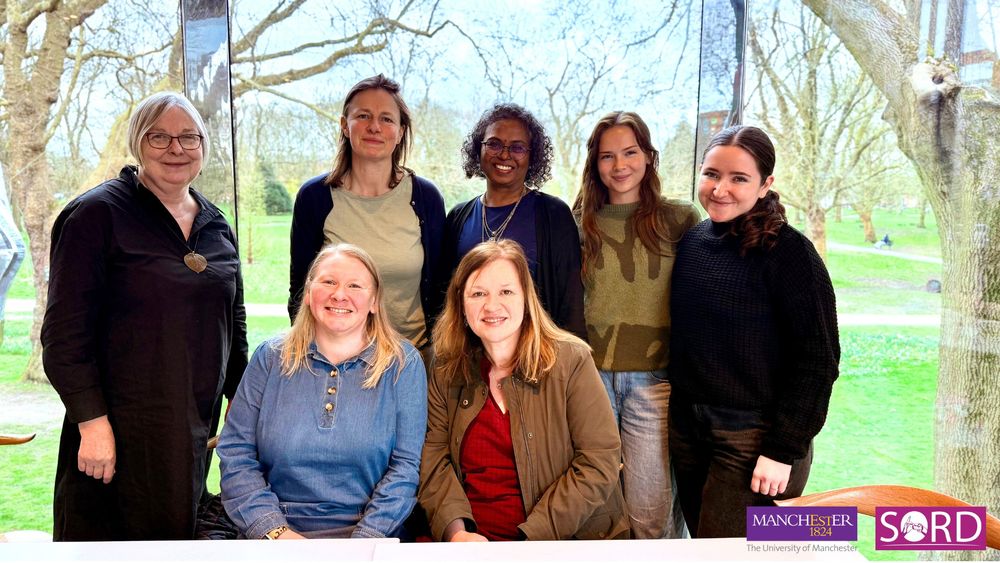 Photo ID: Six people posing together, smiling at the camera inside the café at Whitworth Gallery, Manchester. Four stand in the back row, while two sit in the front. A large window behind them reveals trees and grass. The University of Manchester and SORD logos appear in the bottom right corner.