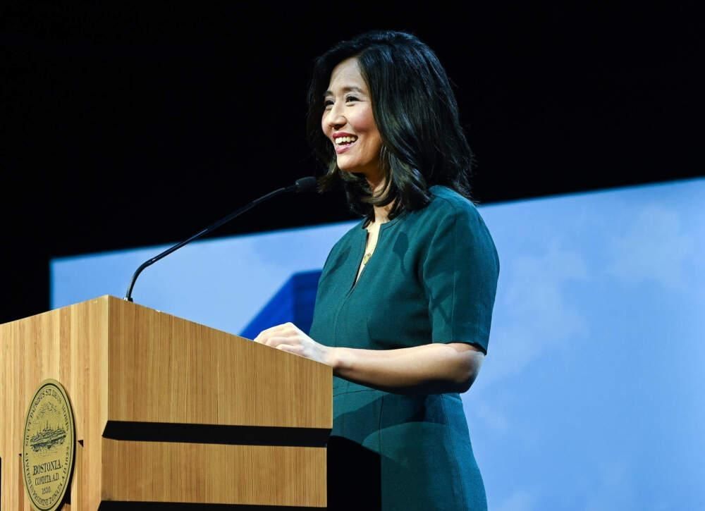 Boston Mayor Michelle Wu smiles at the podium during her State of the City Address. She is wearing green standing in front of a blue backdrop and there is a city of Boston seal on the wooden podium.