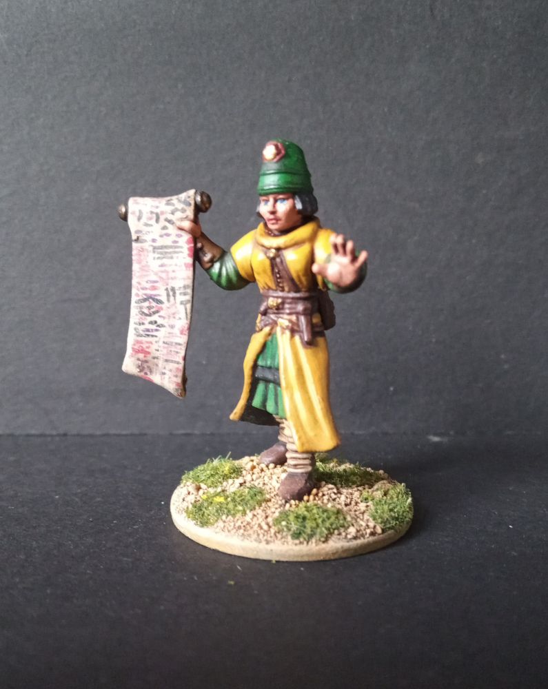 A Frostgrave multi-part plastic wizard, painted wearing a yellow robe and green flowerpot hat, holding a unrolled scroll in her right hand.