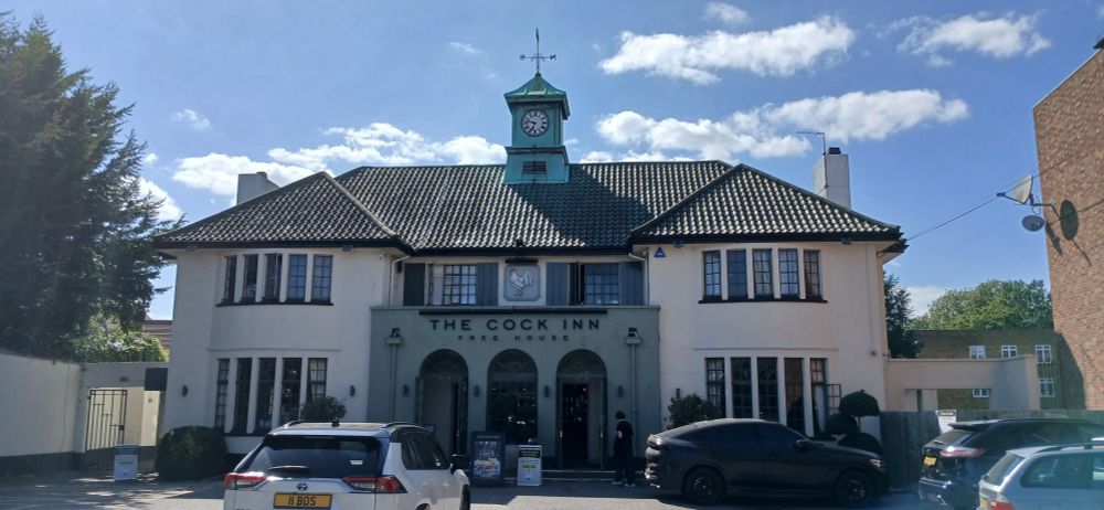 Picture of a white, two storey building with a recessed entryway painted light green announcing "The Cock Inn Free House" above three arches. Above the black tiled roof is a share clock tower with a pointed roof and weather vane.