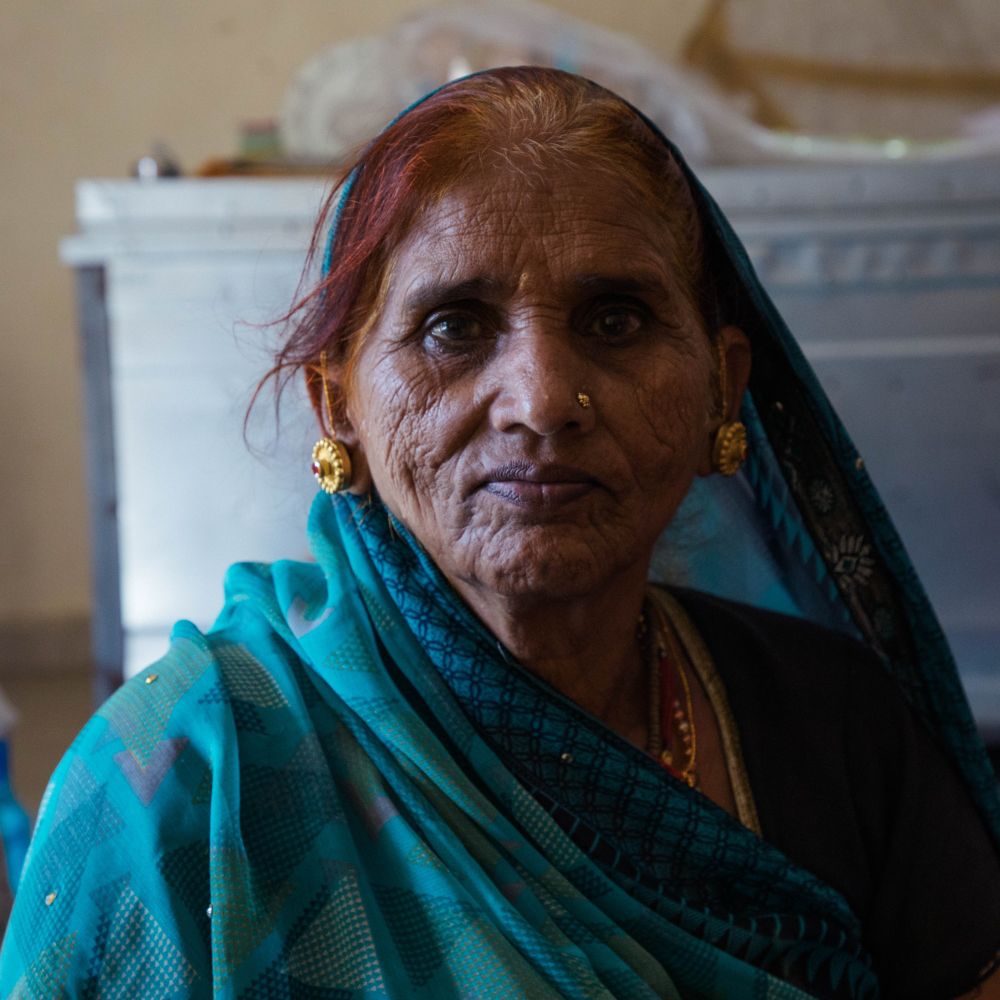 A senior woman in a teal sari wrapped across red dyed hair. She is wearing large gold earrings and has a gold nose stud. She is looking directly at the camera.