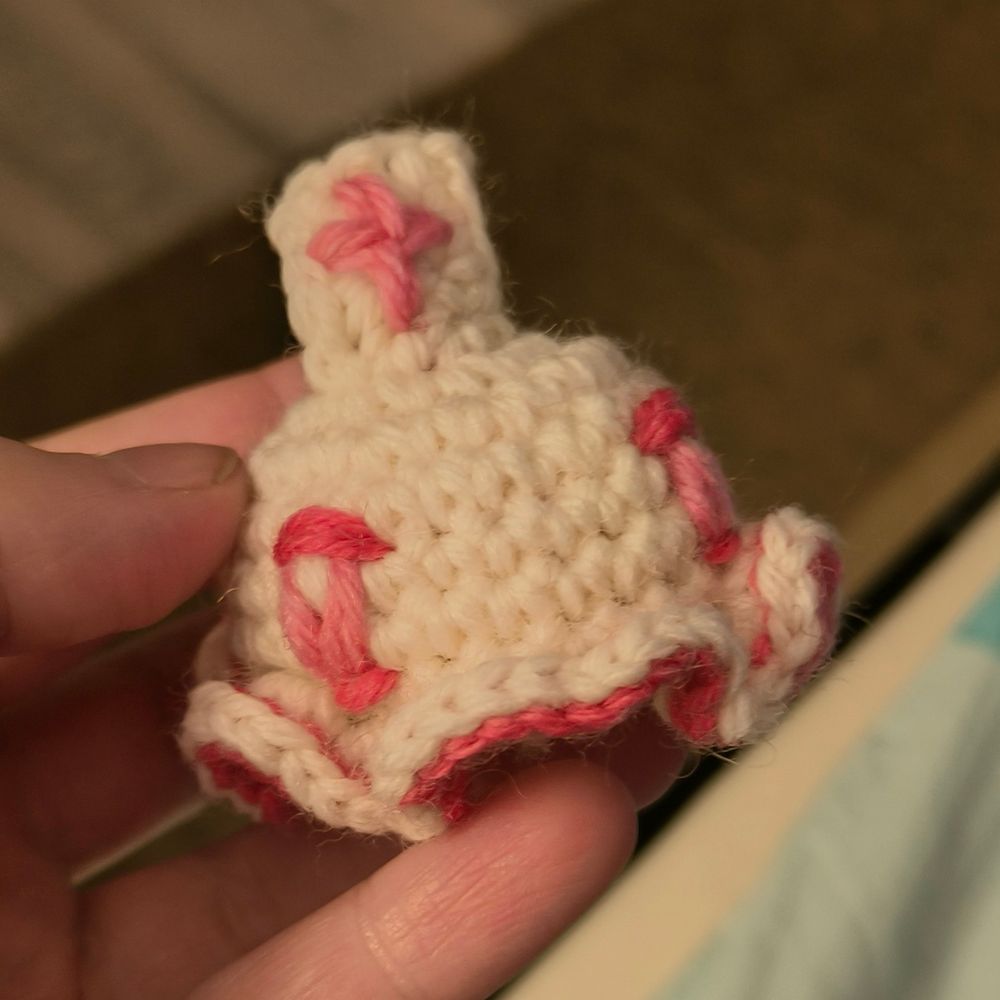 A piss quality picture of a crochet jellyfish. It's white with red and pink accents, wearing a white square with a pink cross on its head.