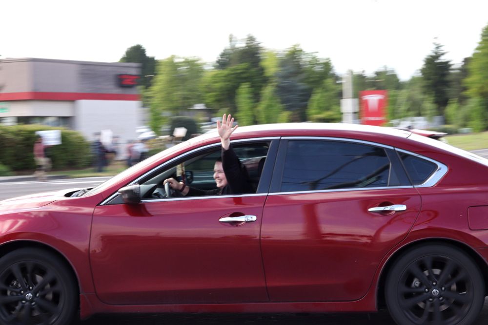 A red car is driving past the viewer. The driver is smiling and waving out of their car window. In the background the sign for the Tesla dealership is visible.

At 5:00pm on May 14th, 2025, protestors gathered outside the Vancouver, WA Tesla dealership to protest the fascist regime of US President Donald Trump and unelected billionaire oligarch Elon Musk. 
