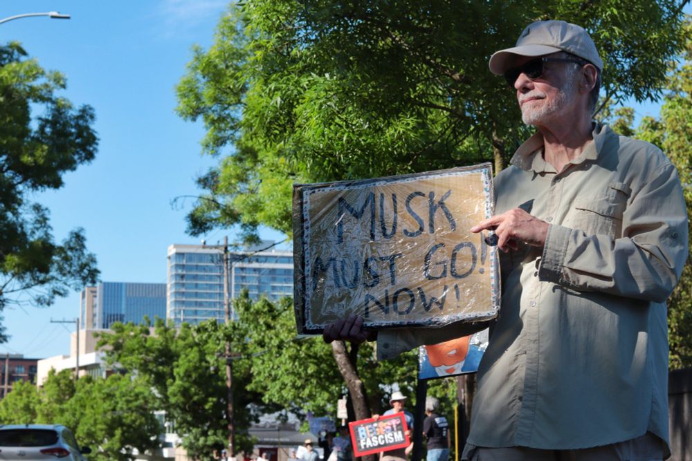 A protestor faces the road holding a picket sign that reads: “MUSK MUST GO! NOW!” The sign is covered in a protective plastic coating. Other protestors can be seen standing on the sidewalk beyond the first protestor. One visible picket sign in the background reads: “RESIST FASCISM”. 

At 3:45pm on May 5th, 2025, protestors gathered outside the Portland Tesla dealership on Macadam Ave to protest the fascist regime of Donald Trump and Elon Musk.