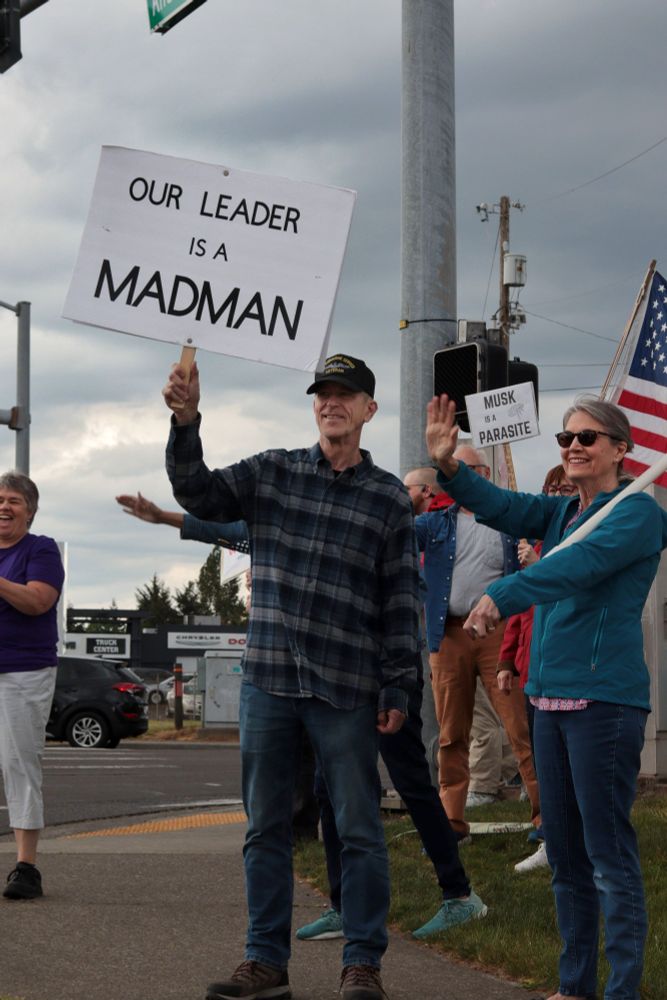 Several protestors stand at the corner of the intersection, smiling at passing cars. One protestor wearing a veteran’s baseball cap lifts a sign that reads: “OUR LEADER IS A MADMAN”. Another protestor holds a sign that reads: “MUSK IS A PARASITE”. A protestor on the right side of the image holds a flag pole, holding a flag that is out of view. Behind them, another protestor holds a US flag. 

At 5:00pm on May 7th, 2025, protestors gathered at the intersection of NE Fourth Plain Blvd and NE Andresen Rd outside the Vancouver, WA, Tesla dealership to protest the fascist regime of President Donald Trump and unelected billionaire Elon Musk.
