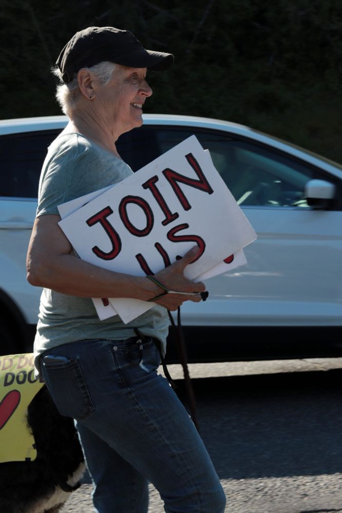 A profile image of a protestor walking to the right in front of the viewer. The protestor is smiling and holding multiple signs in a stack in their right arm. The outermost sign reads: “JOIN US”. A car is visible in the background driving toward the right. 

At 3:45pm on May 5th, 2025, protestors gathered outside the Portland Tesla dealership on Macadam Ave to protest the fascist regime of Donald Trump and Elon Musk.
