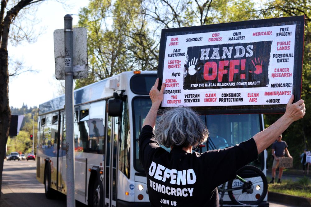 A protestor holds up a picket sign towards a passing city bus. The visible side of the sign reads "HANDS OFF | STOP THE ILLEGAL BILLIONAIRE POWER GRAB!!!" surrounded by a list of essential US social services affected by the Trump Regime, Elon Musk, and DOGE.