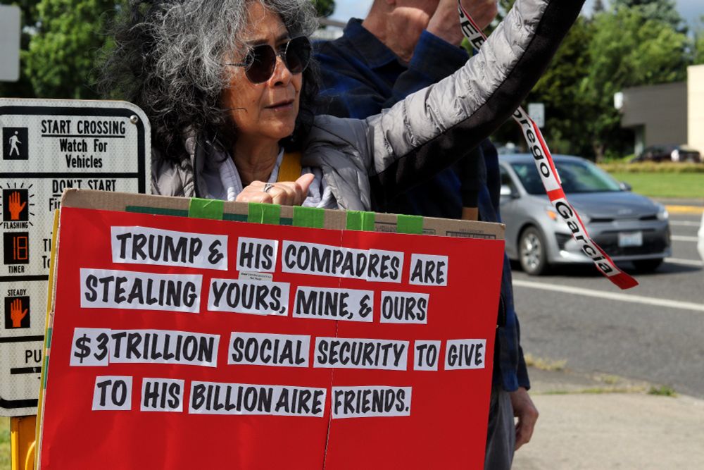 A demonstrator holds up a red picket sign while waving towards passing cars. The visible portion of their sign reads, "TRUMP & HIS COMPADRES ARE STEALING YOURS, MINE, & OURS | $3 TRILLION SOCIAL SECURITY TO GIVE TO HIS BILLIONAIRE FRIENDS."

At 4pm on May 19th, 2025, demonstrators in Vancouver, WA gathered at the intersection of Mill Plain and Chaklov to protest the destruction of essential social services by US President Donald Trump's fascist regime.